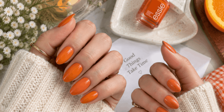 Glossy orange almond-shaped nails on hands styled with cozy cream sleeves, gold rings, fresh flowers, and citrus-inspired beauty accents.